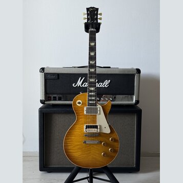 Elektrická kytara typu Les Paul s žlutým plamenovitým topem a bílým pickguardem. Na hlavě hmatníku je zlatý nápis Gibson. Kytara stojí na stojanu pře…