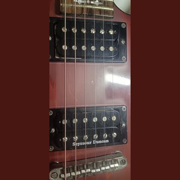 Detail korpusu červené elektrické kytary s dvěma černými humbuckery. Na spodním snímači je bílý nápis Seymour Duncan. Povrch laku vykazuje drobné škr…