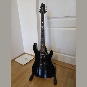 Černá elektrická kytara s dvojsekmem, umístěná na černém stojanu. Na hlavě nástroje je bílý nápis CORT. Kytara má dva humbuckery a dva ovládací knofl…