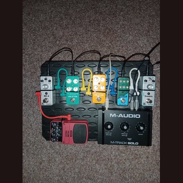 Na černém efektovém pedalboardu je umístěno šest kytarových pedálů a jedno audio rozhraní. Zleva doprava jsou řazeny pedály s nápisy AD-6, VALETON TU…