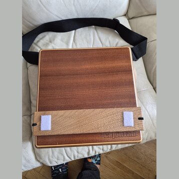 Dřevěný cajon s hnědým povrchem a světlými lemovými lištami. V dolní části je vyryt nápis „CajonLab“. Nástroj je doplněn o světlý dřevěný příslušenko…