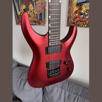 Elektrická kytara s červeným matným povrchem a hnědým hmatníkem. Nástroj má dva černé humbuckery a černý most s integrovaným pákovým systémem. Na těl…