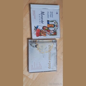 Dvě CD v plastových obalech položená na dřevěném povrchu. Horní obal má nápis „Moravské koledy“ a jména „Bambini di Praga z Hradistana“, „Bohumil Kul…