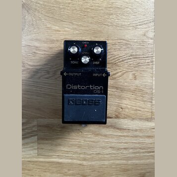 Černý efekt pedál s nápisy Distortion a DS-1. Na horní části jsou tři stříbrné ovládací knoflíky s popisky LEVEL, TONE a DIST, červená kontrolka CHEC…