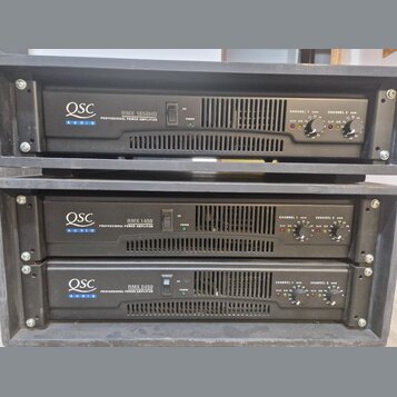Tři černé rackové výkonové zesilovače značky QSC AUDIO. Horní jednotka nese nápis RMX 1850HD PROFESSIONAL POWER AMPLIFIER, prostřední RMX 1450 PROFES…