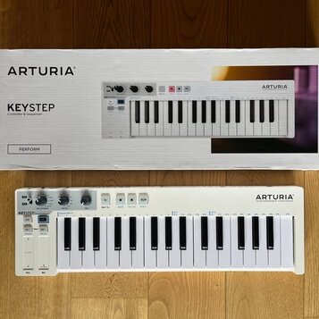Bílá klávesová kontrola s logem ARTURIA a názvem KEYSTEP. Nástroj má plastové tělo, bílé klávesy a ovládací prvky v horní části. Vedle nástroje je um…