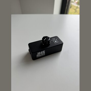 Černý efekt pedál obdélníkového tvaru s jedním ovládacím knoflíkem. Na boku je bílý nápis JHS PEDALS a pod ním menší text LITTLE BLACK AMP BOX. V hor…