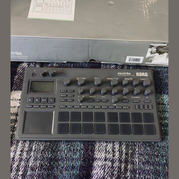 Šedý sampler/sekvencer značky KORG s nápisem electribe MUSIC PRODUCTION STATION. Zařízení obsahuje několik otočných potenciometrů, digitální displej…