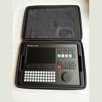 Černý sampler Polyend Tracker v černém přepravním pouzdře. Zařízení má displej, mřížku bílých tlačítek a několik barevných ovládacích prvků s nápisy…