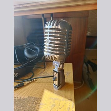 Stříbrný mikrofon s kovovou mřížkou v retro stylu, umístěný na stolním stojanu. Na přední straně mřížky je viditelné kulaté logo s písmenem „S“. Na s…