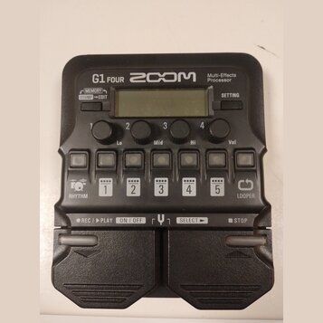Černý multi-efektový procesor s nápisy G1 FOUR, ZOOM a Multi-Effects Processor. Zařízení obsahuje displej, čtyři otočné ovladače s popisky Lo, Mid, H…