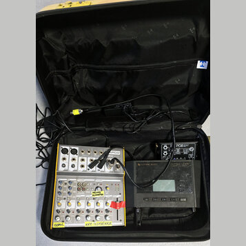 Černý přenosný kufr s elektronickým vybavením. Uvnitř jsou dva mixážní pulty značky TAPCO, z nichuž horní nese nápis MIX 600. Na spodním mixážním pul…