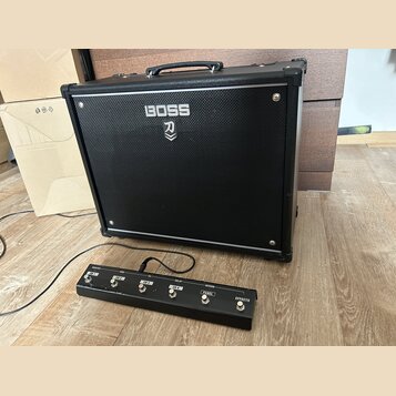 Černý kytarový kombozesilovač s logem BOSS a pod ním symbolem stylizovaného meče. Před zesilovačem je umístěn černý nožní spínač s nápisy BOOSTER, MI…