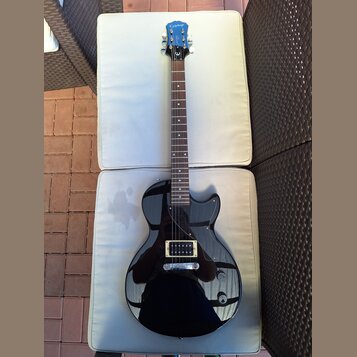 Černá elektrická kytara s tmavým hnedým hmatníkem a černým pickguardem. Na hlavě je bílý nápis Epiphone. Nástroj vykazuje známky běžného používání, p…