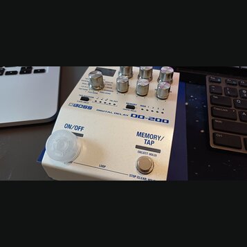 Bílý efekt pedál s nápisy BOSS DIGITAL DELAY DD-200. Na horní části jsou ovládací knoflíky a přepínače s popisy funkcí, jako je TAP DIVISION nebo MEM…