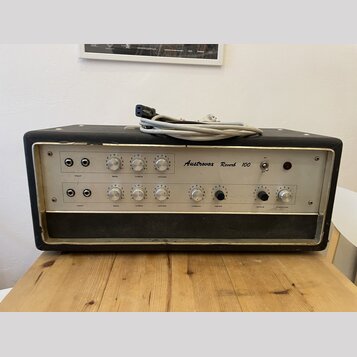 Černý kytarový kombozesilovač s bílým předním panelem a kovovým rámem. Na panelu je nápis Austrovox Reverb 100 a řada ovládacích knoflíků s popisky i…