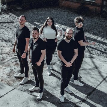 Fotografie zobrazuje skupinu pěti lidí stojících venku na betonovém povrchu. Čtyři muži mají na sobě černé trička a černé kalhoty, jedna žena je oble…