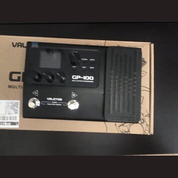 Černý kytarový procesor s integrovaným pedálem, umístěný na kartonové krabici. Na zařízení je uveden nápis GP-100 a značka VALETON. Jsou vidět ovláda…