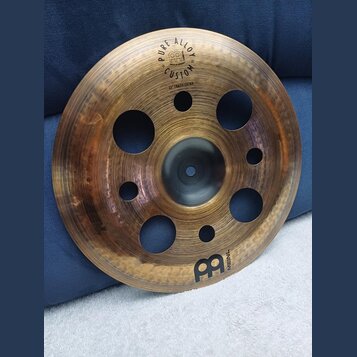 Kruhový činel z mosazného kovu s několika otvory různých velikostí v ploše. Na povrchu jsou nápisy PURE ALLOY, CUSTOM a 12" TRASH CHINA spolu s logem…