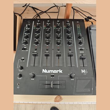 Černý mixážní pult s nápisem Numark a označením M6 USB. Zařízení obsahuje čtyři vstupní kanály (CH1 až CH4) a jeden Master kanál, každý s posuvníkem…