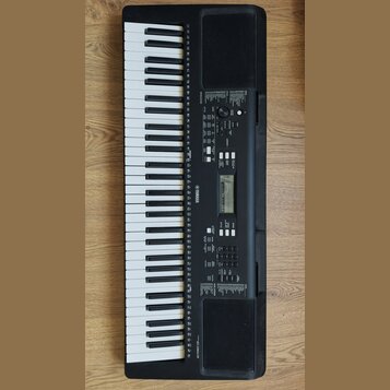 Černý digitální klavír s bílými a černými klávesami. Na korpusu je vlevo nápis PSR-E323 a uprostřed logo YAMAHA. Nástroj obsahuje ovládací panel s di…