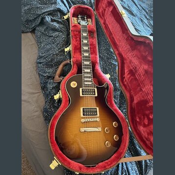 Elektrická kytara s trupem ve tvaru Les Paul v barevné variantě sunburst, umístěná v otevřeném tvrdém kufru s červeným polstrováním. Nástroj má dva h…