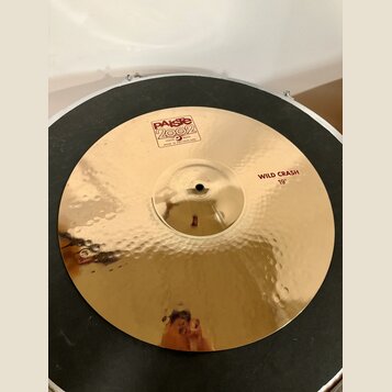 Zlatý kovový činel s kladeným povrchem. Na povrchu jsou červené nápisy „Paiste 2002“, „TRADE MARK MADE IN SWITZERLAND“ a „WILD CRASH 19"“. Povrch je…
