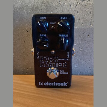 Černý efekt pedál s nápisy GAIN, LEVEL, BASS a TREBLE u ovládacích knoflíků a přepínačem VOICE. Uprostřed je velký bílý nápis DARK MATTER DISTORTION…