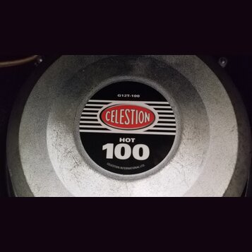 Detailní pohled na střed reproduktoru s kovovým šedým krytem. Uprostřed je černý štít s červeným logem CELESTION a bílými nápisy HOT 100, G12T-100 a…