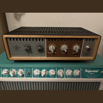 Na fotografii je stolní audio zařízení s černým perforovaným krytem a dřevěným rámem. Na levé straně je logo UA a nápis UNIVERSAL AUDIO, vpravo nápis…
