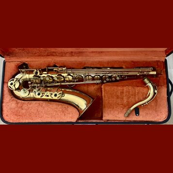 Zlatý saxofon v otevřeném černém kufru s červeným polstrováním. Nástroj je v rozloženém stavu, vedle něj leží samostatně hnedbek. Na těle nástroje ne…