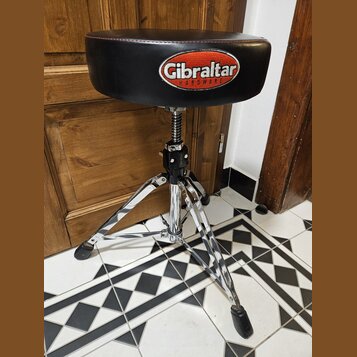 Černý polstrovaný bubenícký židun s chromovaným třínohým stativem. Na sedadle je bílo-červené logo s nápisem Gibraltar HARDWARE. Na černé části mecha…
