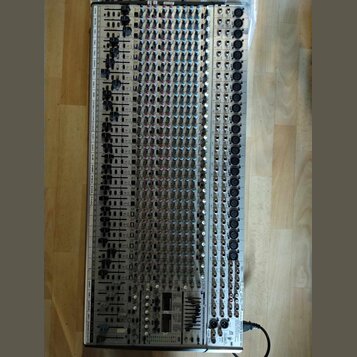 Stříbrný mixážní pult s velkým množstvím regulačních knoflíků, posuvníků a vstupních konektorů. Zařízení je umístěno na dřevěné podlaze. Na spodním p…