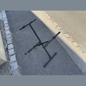 Černý kovový stojan na hudební nástroj s dvěma polstrovanými oporami. Stojan má sklapovací konstrukci a stojí na asfaltovém povrchu. Na fotce nejsou…