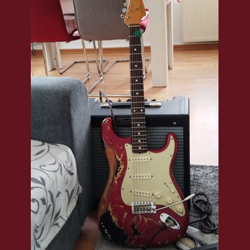 Elektrická kytara typu Stratocaster v červené barvě s výrazně odřeným povrchem a viditelným dřevem. Má krémový pickguard, tři snímače a chromovaný mo…