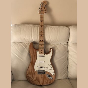 Elektrická kytara s přírodním dřevěným povrchem těla a hmatníkem. Na hlavě je viditelné logo Fender Stratocaster. Bílý pickguard obsahuje tři snímače…