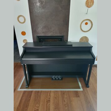 Černý digitální piano v kompaktním provedení s integrovaným stojanem. Nástroj má matný povrch a v dolní části jsou umístěny tři pedály. Na viditelnýc…