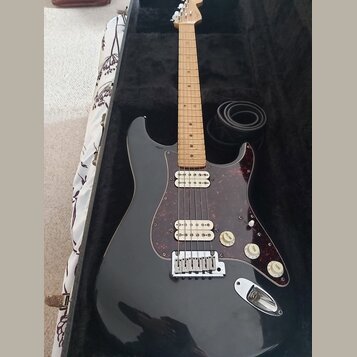 Černá elektrická kytara s tvarem typu Stratocaster, světlým hmatníkem a červeně vzorovaným pickguardem. Nástroj má tři snímače, tři ovládací knofíky…