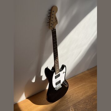 Černá elektrická kytara s bílým pickguardem a hnědým hmatníkem. Na hlavě nástroje je viditelné logo Squier. Kytara je opřena o bílou zeď na dřevěné p…