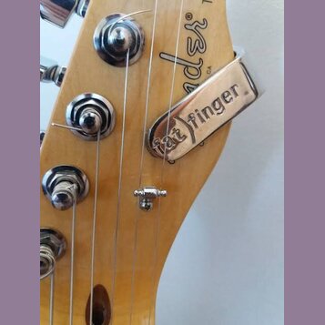 Detail hlavy elektrické kytary z přírodního dřeva. Na dřevě je viditelné částečné logo Fender. Na strunách je upevněn kovový stahovací kousek s nápis…