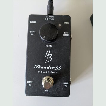 Černý výkonový zesilovač v kovovém boxu s nápisy Thunder 99 a POWER AMP. Uprostřed je logo HB a ovratový knoflík s popiskem VOLUME. Zařízení má vstup…