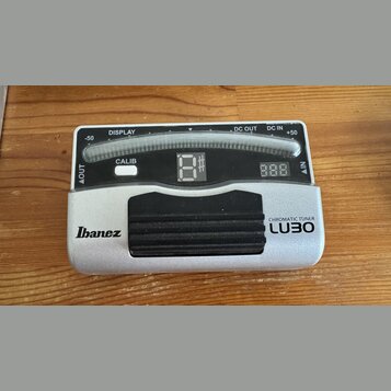 Stříbrný chromatický tuner s černým gumovým pedálem. Na zařízení jsou nápisy Ibanez, CHROMATIC TUNER a LUBO. Horní část obsahuje displej s označením…