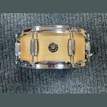 Fotografie znázorňuje snare drum s dřevěným korpusem v přírodní světle barvě a chromovaným obručemi. Uprostřed korpusu je umístěno kulaté logo s nápi…