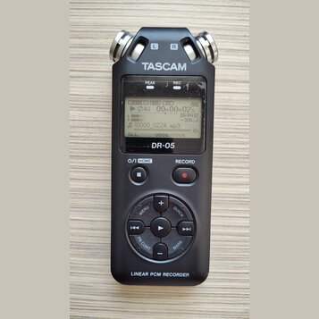 Černý digitální záznamový systém s dvěma stříbrnými mikrofony v horní části označenými písmeny L a R. Na přístroji je bílý nápis TASCAM, modelové ozn…