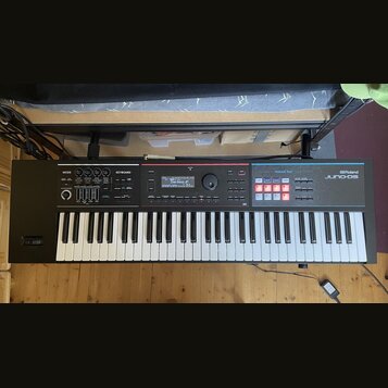 Černý digitální klavír s bílými a černými klávesami. Na pravé straně je nápis Roland JUNO-DS a modrá sekce s označením PHRASE PAD. V horní části jsou…