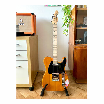 Elektrická kytara typu Telecaster v žluté barvě s černým pickguardem a světlým hnedým dřevěným hmatníkem. Na hlavě je vidět nápis "Squier" a pod ním…