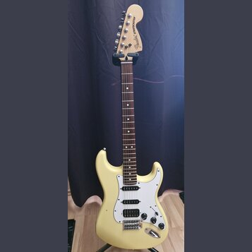 Elektrická kytara s krémovým korpusem, bílým pickguardem a hnedou hmatníkovou deskou. Na hlavě je černý nápis „Fender STRATOCASTER“. Nástroj má tři s…