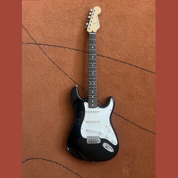 Černá elektrická kytara s bílým pickguardem a hnedým hmatníkem. Na hlavě je viditelné logo Fender. Nástroj má tři snímače typu single-coil a chromova…