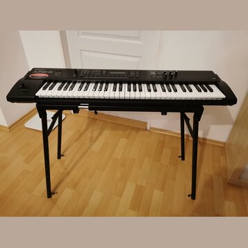 Černý digitální klavír umístěný na černém X-stojanu. Na levé straně korpusu je bílý nápis Roland a pod ním modelové označení Juno-D, doplněný červený…