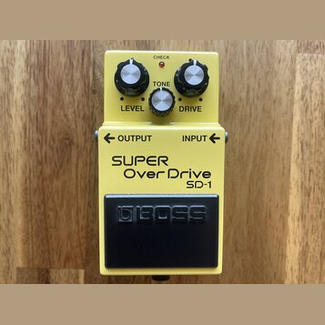 Žlutý efekt pedál s černým spínacím tlačítkem. Na spodní části je nápis SUPER Over Drive SD-1 a na spínači logo BOSS. V horní části jsou tři ovládací…